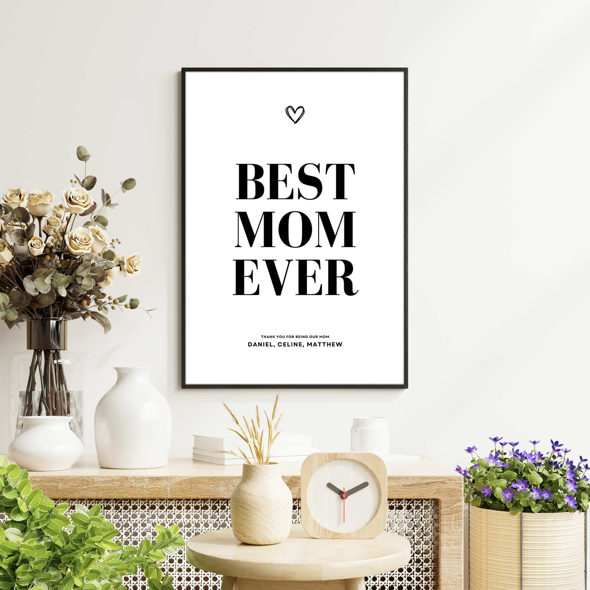 Framed ‘Best Mom Ever’ poster hanging above a wooden sideboard decorated with vases, flowers, and plants in a bright modern living room.
